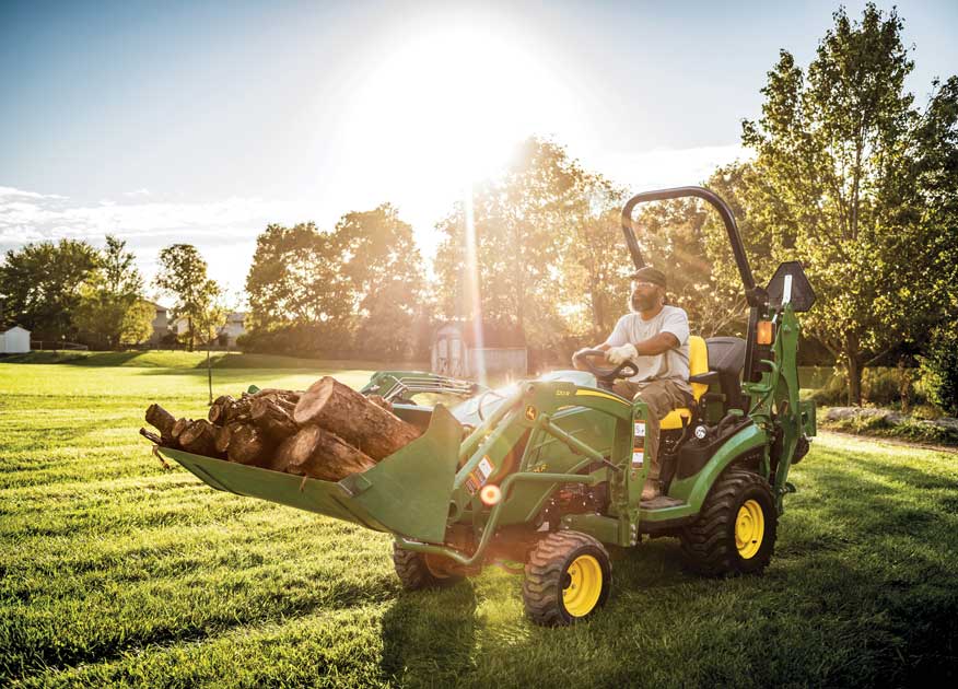 man operating a John Deere 1025R Compact Tractor with a 120R bucket attachment with logs in it