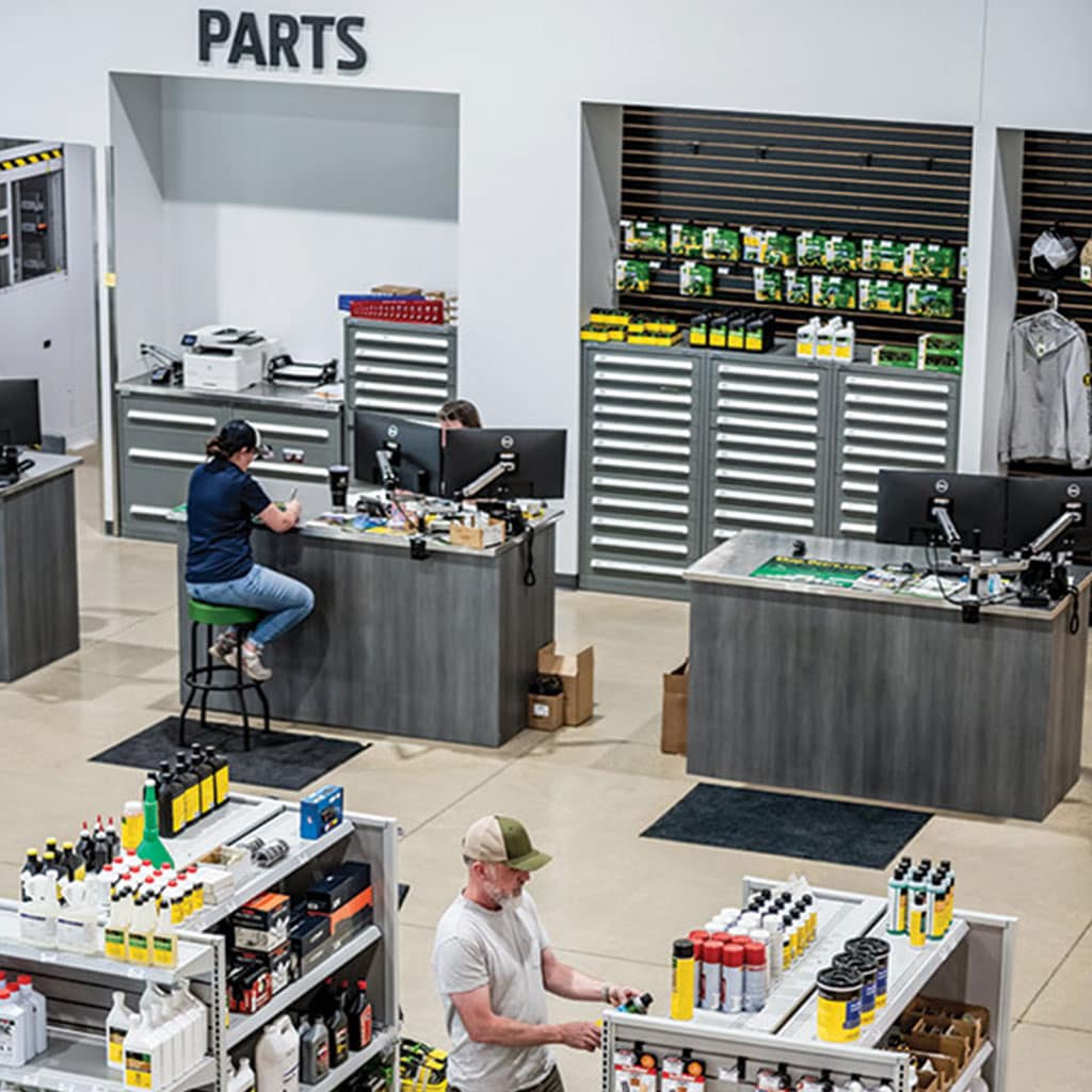 Two staff members assist in parts showroom with workstations and shelves stocked with automotive products.