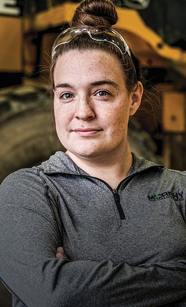 Michelle Roth is standing in a Murphy Tractor service bay with a John Deere 844K Loader behind her while looking at the camera.