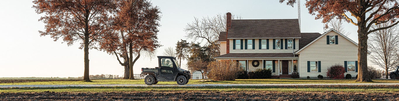 Gator™ XUV835M UTV driving in front of a white house. Two people are in the front seat.