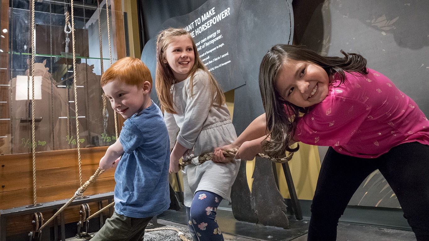 Trois jeunes enfants tirant des cordes sur un affichage interactif et découvrant combien il est difficile de tirer une charrue