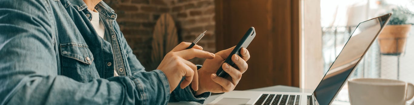 Man sitting at a desk with a laptop open and using his cell phone with a pen in his hand