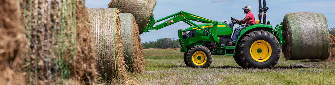 Tracteur compact de John Deere muni d'une fourche en train de soulever une balle de foin dans le champ