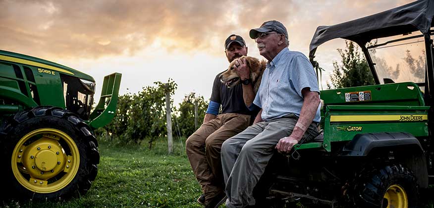Deux hommes assis dans la partie arrière d'un tracteur Gator™ de John Deere avec un golden retriever