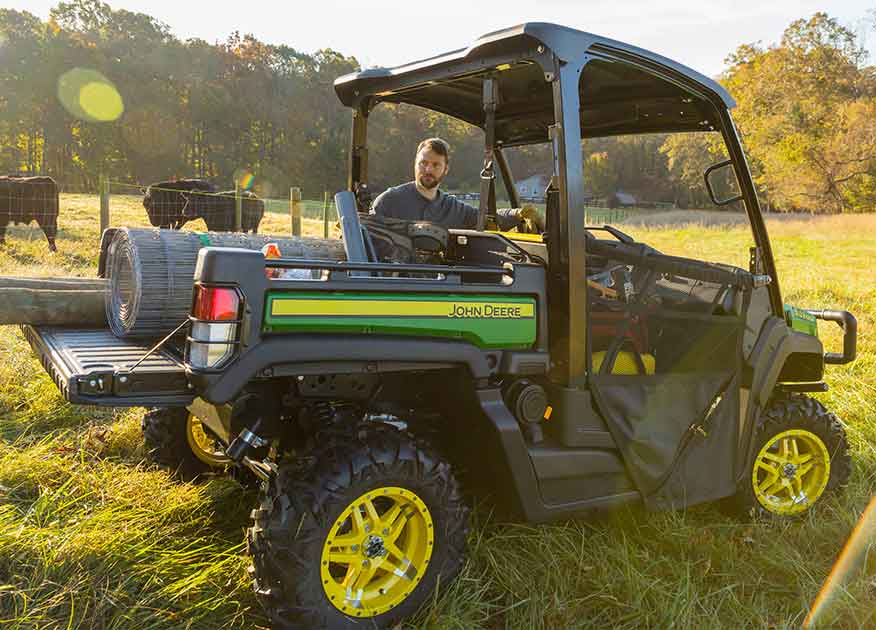 Homme à côté d'un véhicule utilitaire Gator de John Deere qui transporte de l'équipement dans sa caisse de chargement avec du bétail en arrière-plan