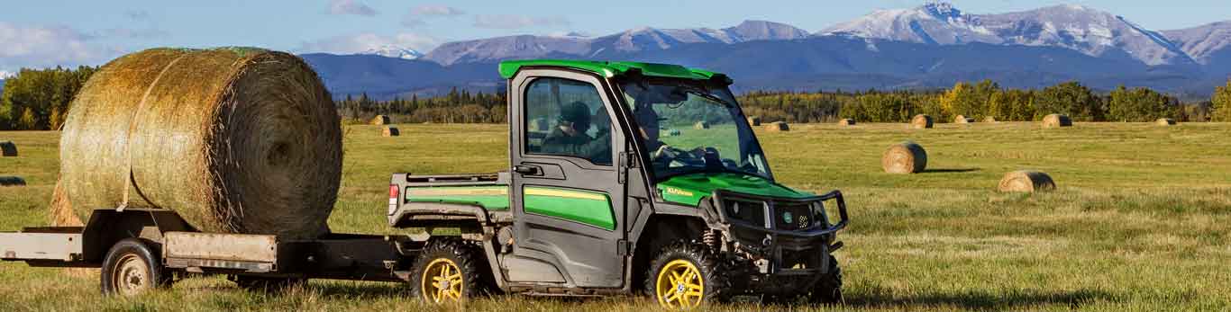 Deux personnes traversant un champ à bord d'un véhicule utilitaire Gator de John Deere, et balle de foin sur une remorque