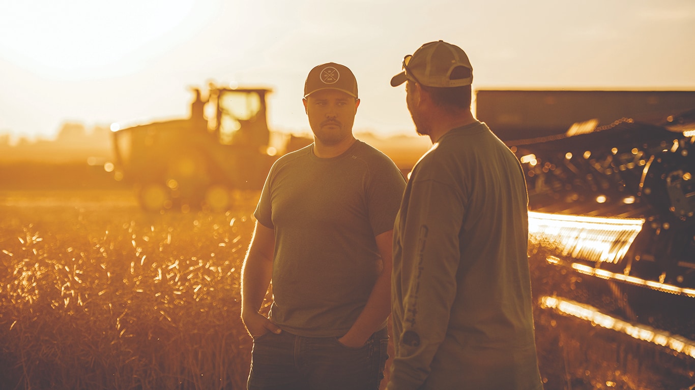 Two people standing in a field at sunset, talking near farming equipment and a tractor in the background.