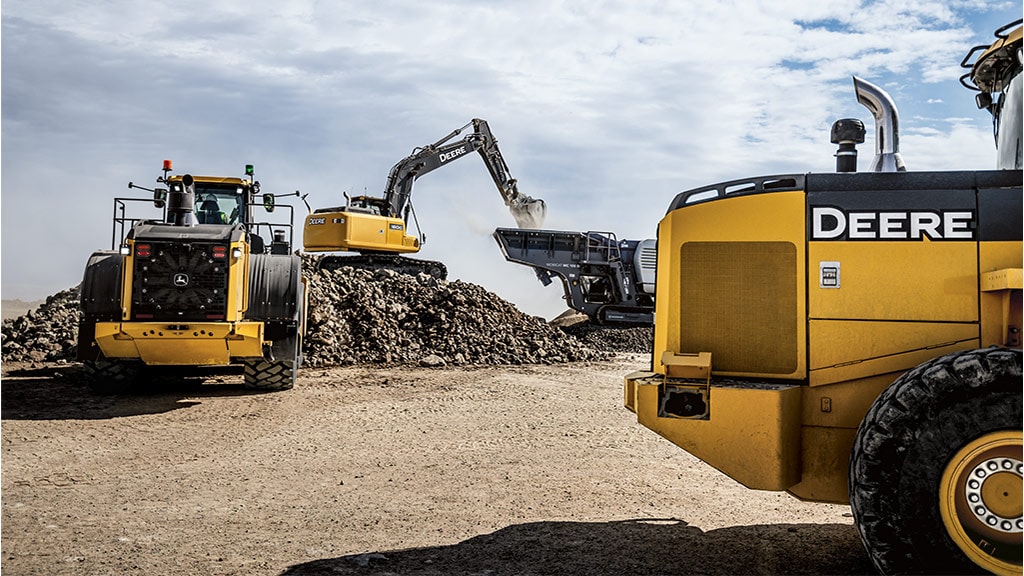 Excavatrice John Deere chargeant de la roche dans un concasseur à côté d’un équipement lourd dans une carrière.