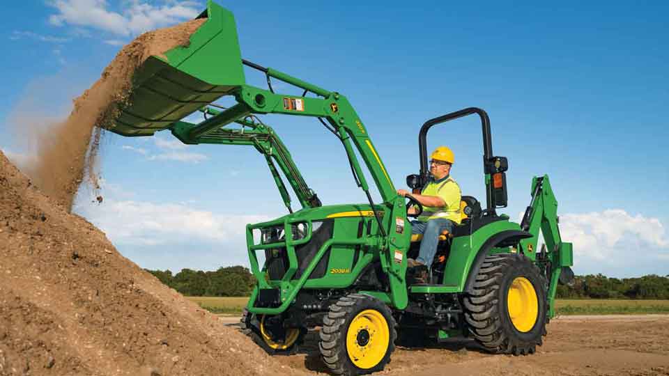 Homme portant un gilet jaune et un casque de sécurité qui déverse de la terre avec un tracteur compact 2038R de John Deere doté d'un godet