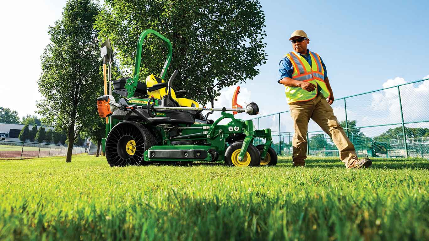 Homme portant une veste jaune qui marche près d'une tondeuse à rayon de braquage nul de John Deere dotée d'un taille-bordure et d'une pelle