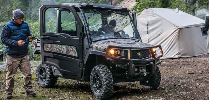 Camouflage John Deere Gator Utility Vehicle in a wooded area with one man inside and another standing outside and a tent in the background