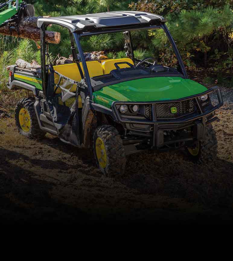 John Deere 835M Crossover Gator Utility Vehicle in a field with wood being loaded in the back by a compact tractor attachment