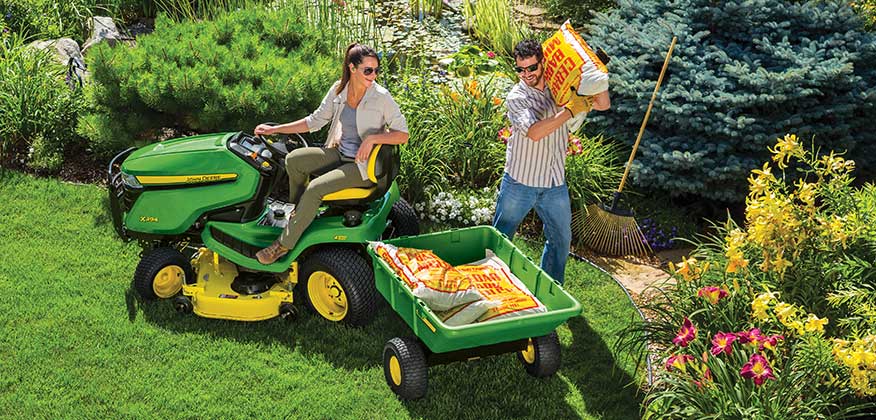 Woman sitting on a John Deere Lawn Tractor in a yard with a man loading fertilizer to the attached poly cart