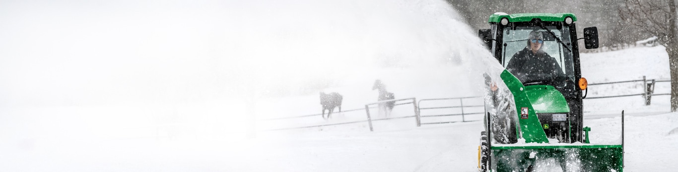 A woman using a John Deere 1025R Sub-Compact Tractor with a snow blower attachment to clear snow from her property.
