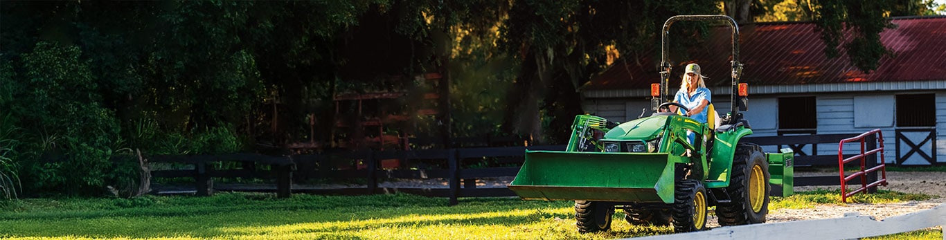 A woman driving a John Deere 3E Series Tractor on a rural property.