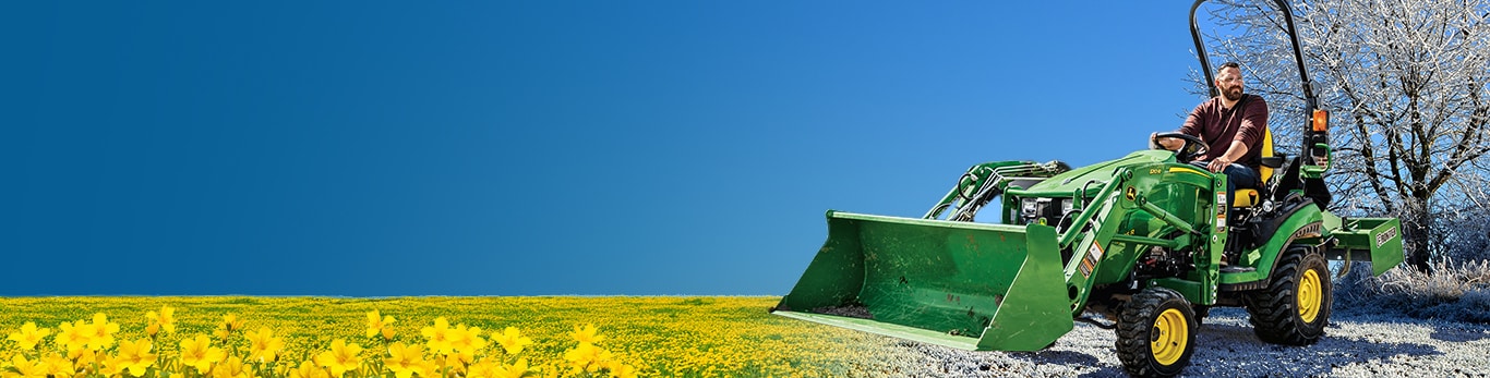 A man driving a John Deere 1025R Compact Tractor with loader attachment from snow-covered grass to blooming fields.