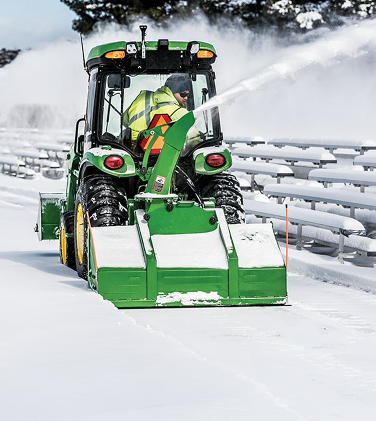 A man using a John Deere 1025R Sub-Compact Tractor to clear snow from a stadium.
