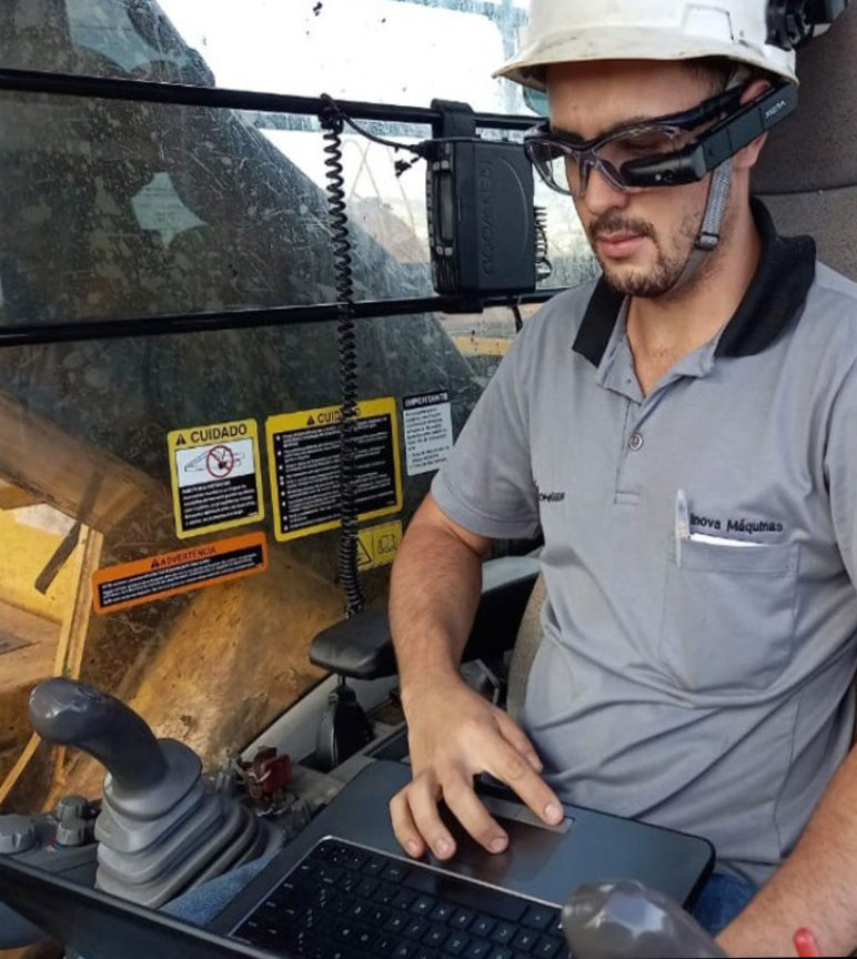 A man in a grey t-shirt wearing a white hard hat using a computer wearing glasses with a black attachment.