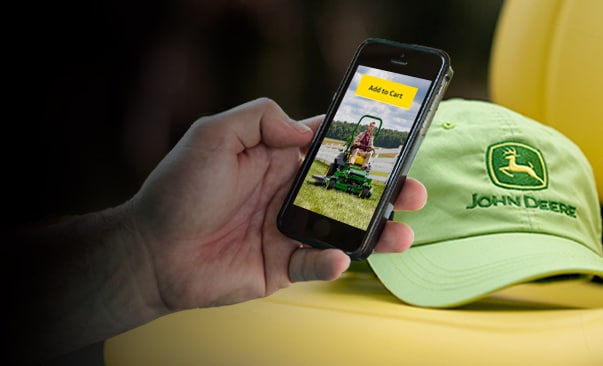 Hand holding a mobile phone, screen has 'Add to Cart' button. John Deere hat in background on yellow seat.