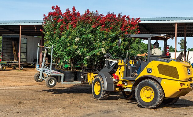 Une chargeuse compacte à roues 184 de catégorie G de John Deere soulève une palette d'arbres en fleurs pour un aménagement paysager.
