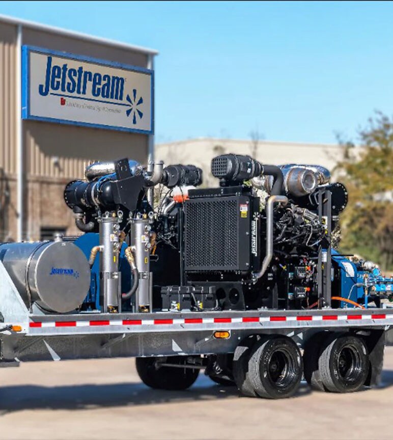 A big blue, silver, and black machine with pipes going through it on the back of a trailer with a Jet Stream Sign in the background.