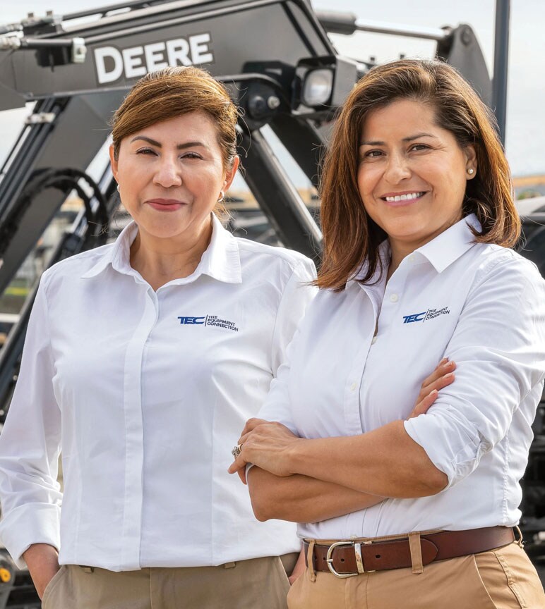 Lilia Jubrail and Ernestina Rincon stand in front of a John&nbsp;Deere excavator.