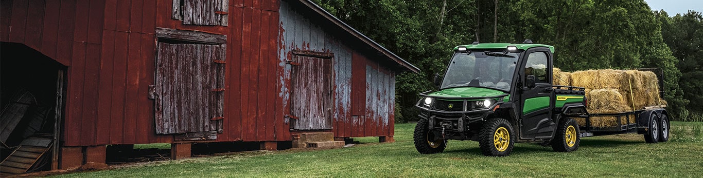 A John Deere Gator™ XUV 845R Signature Edition utility vehicle hauling hay bales by a red barn.