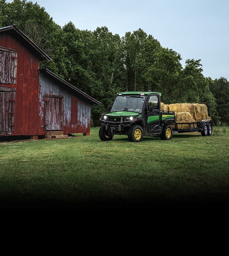 Gator with trailer full of hay bales next to a worn red barn.