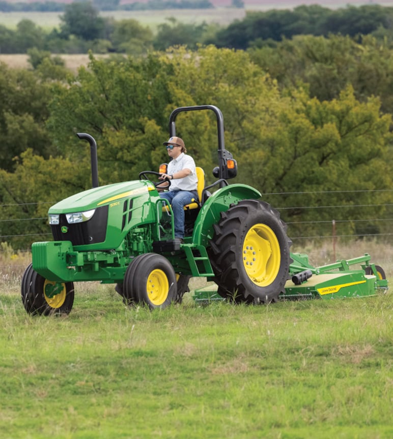 Un homme qui utilise un tracteur utilitaire à plate-forme de conduite John Deere 5050E avec un outil de coupe rotatif pour tondre l’herbe.