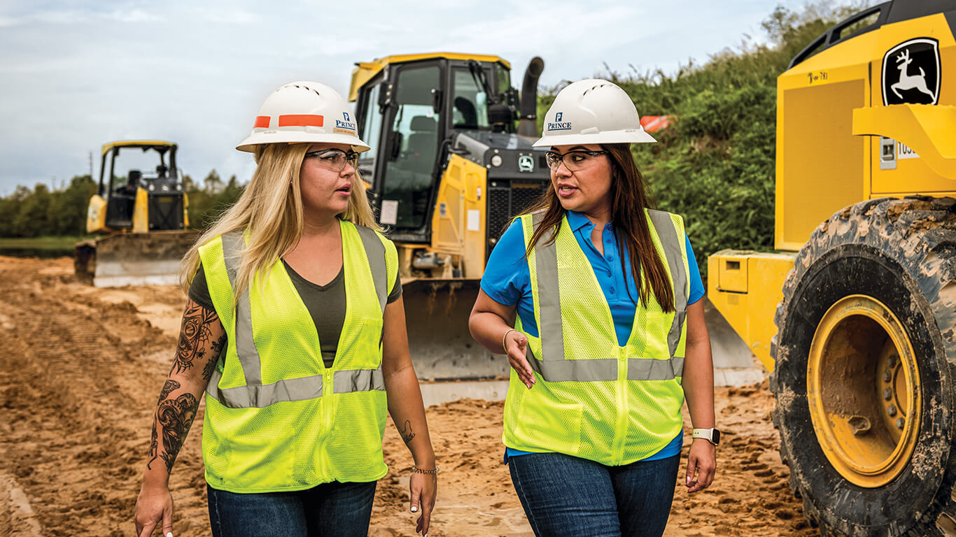 Image de deux femmes portant des casques durs, des lunettes de sécurité et des gilets de sécurité, marchant sur le chantier avec des machines John Deere en arrière-plan.