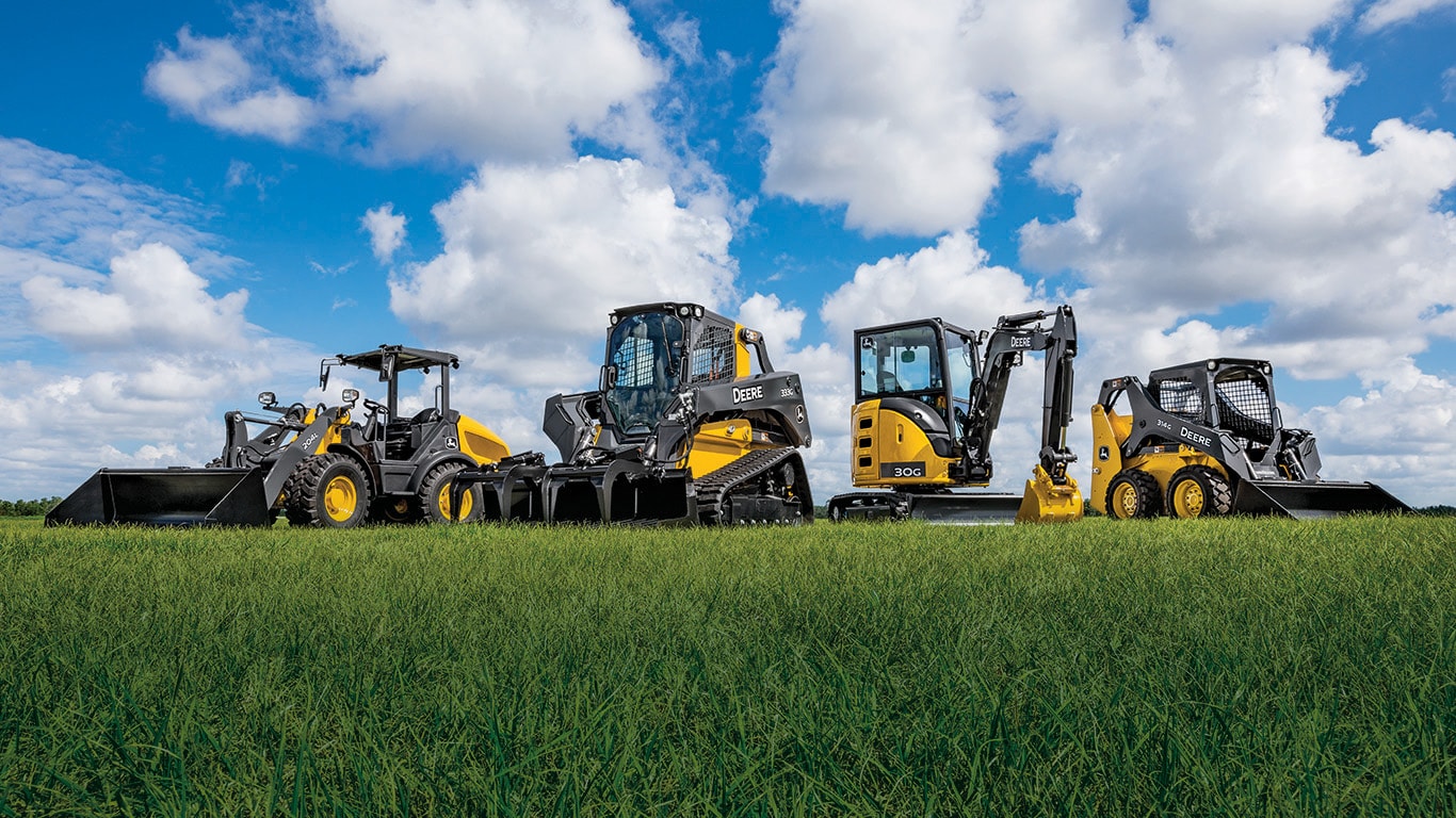 De nombreux équipements de construction compacte John Deere sont garés dans un champ d'herbe.