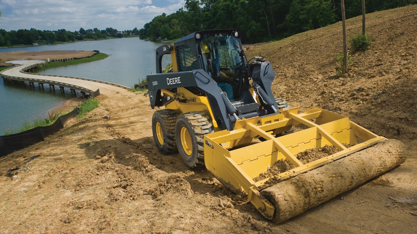 John Deere Skid Steer with vibratory roller attachment flattening dirt near a lake.