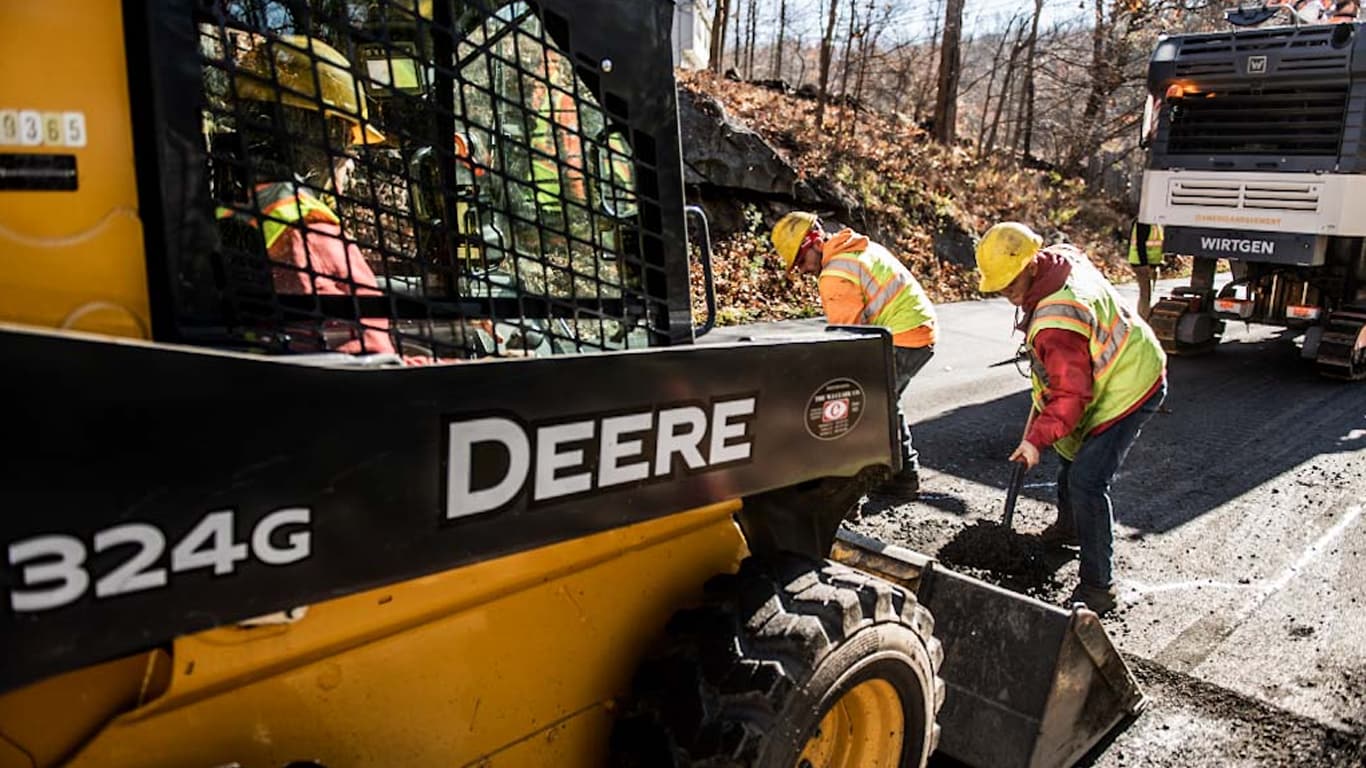 Two men shoveling dirt behind a 324G with a Wirtgen Compact Milling Machine in the background.