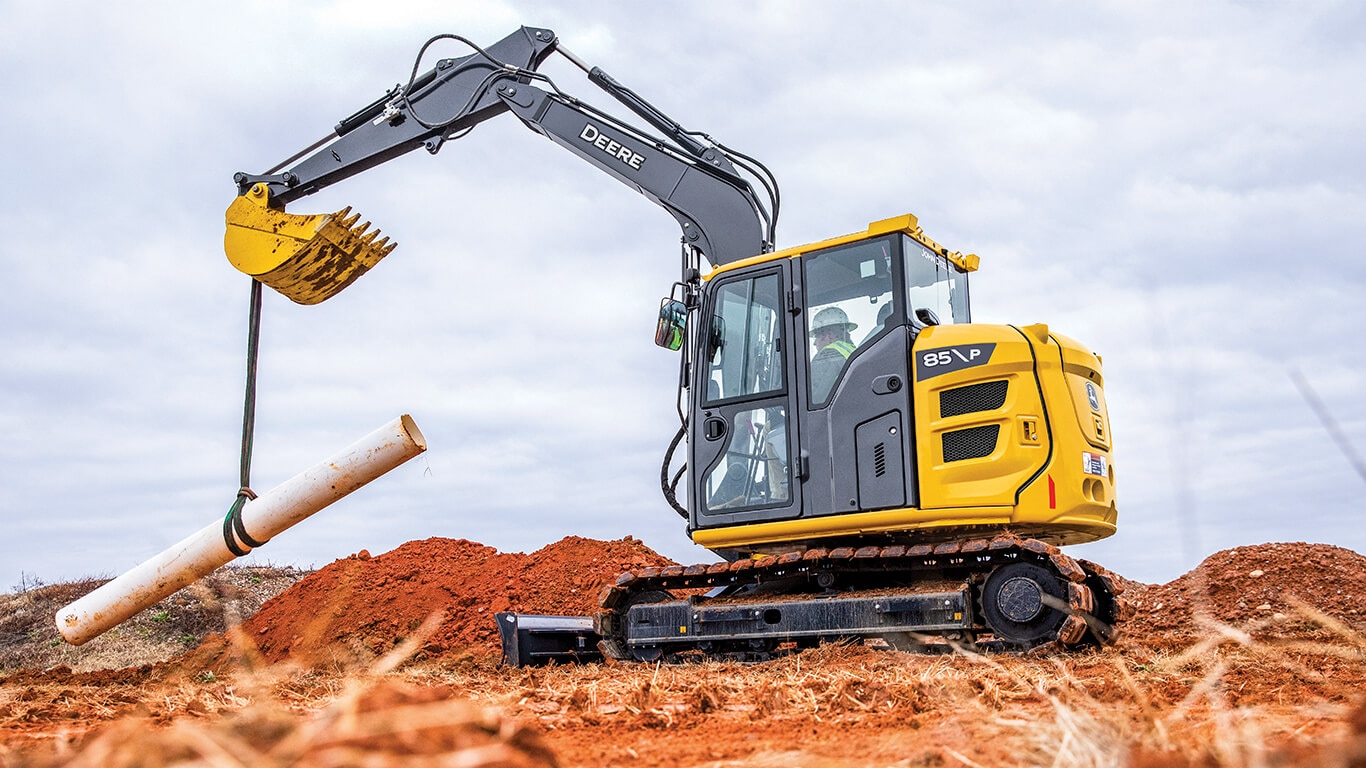 Excavatrice 85 de catégorie P soulevant un tuyau sur un chantier.