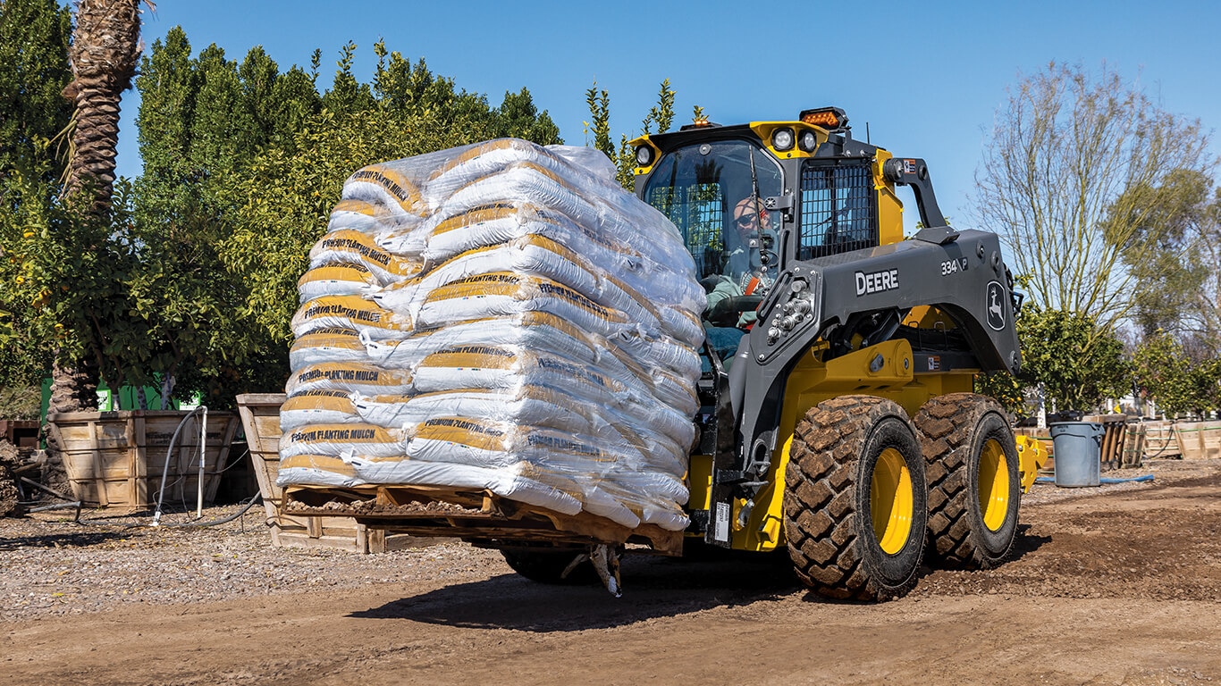 334 P-Tier skid steer moving a pallet of mulch with Deere fork attachment.