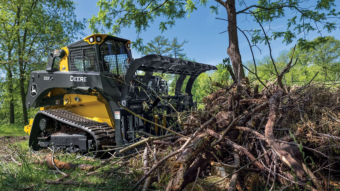 333 P-Tier compact track loader using a John Deere rake attachment to move brush pile of branches and twigs