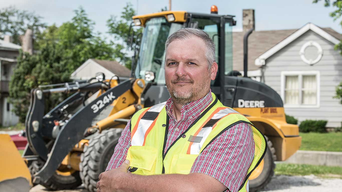 Jessie Plant from Plant Brothers Excavating poses with crossed arms in front of a 324K Wheel Loader on a residential street