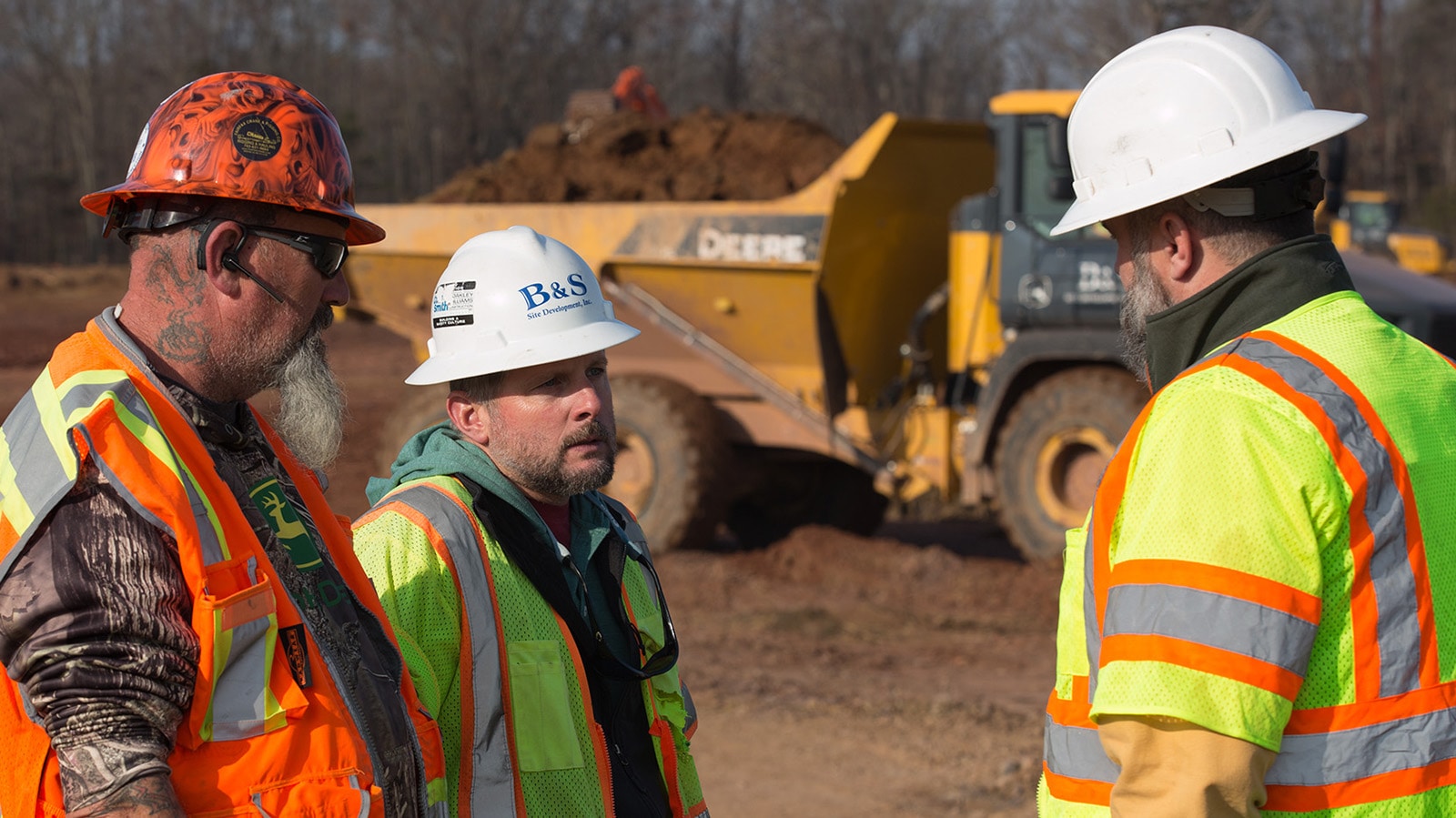 Three people on a jobsite talking with a dump truck full of dirt in the background