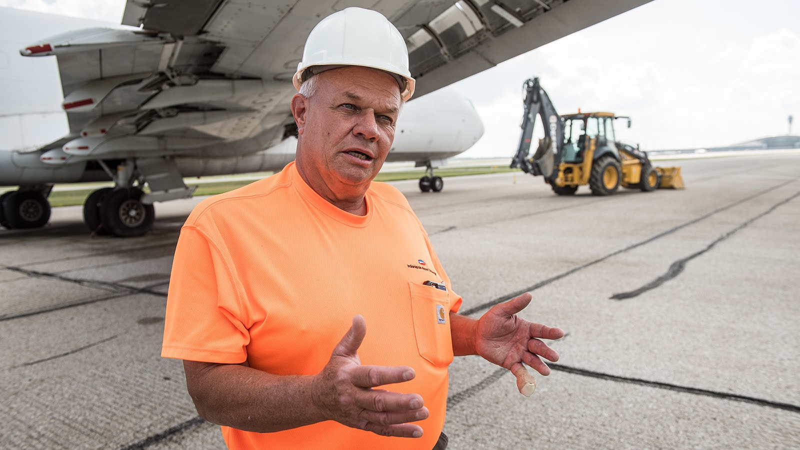 George Kestler stands under the wing of a plane with the 710L Backhoe in the background
