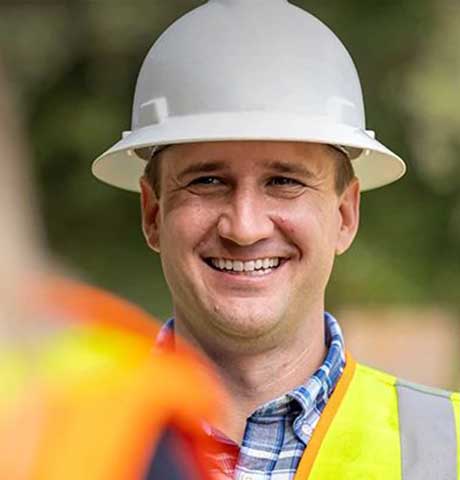 Worker in hard hat and yellow vest stands outdoors; face blurred, another in orange vest nearby.