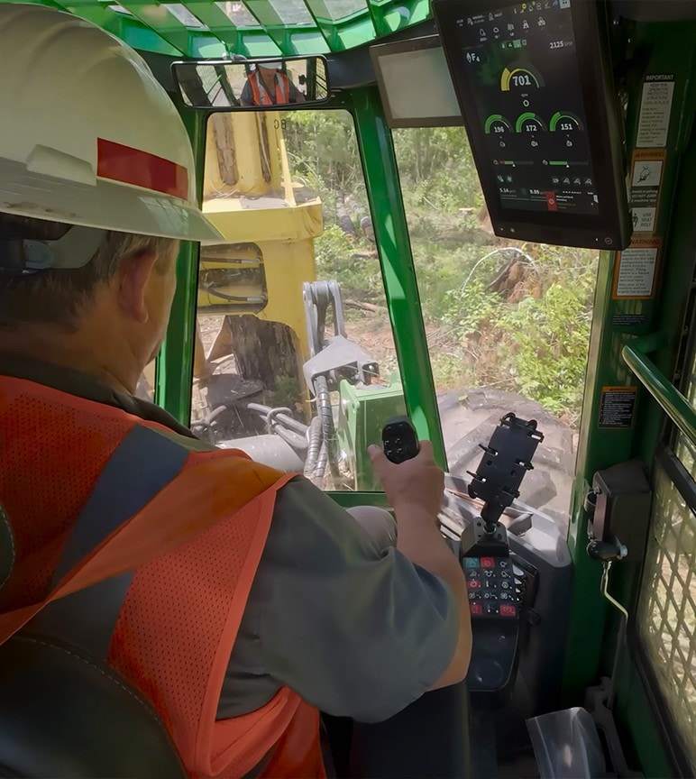 Operator inside forestry machine cab using joystick with digital display showing controls.
