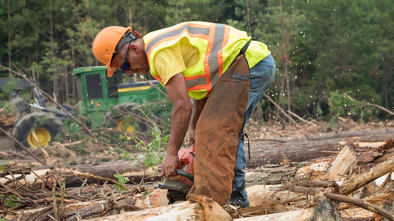 A logger in hard hat, safety glasses and vest operates a chain saw while a John Deere machine works in the background