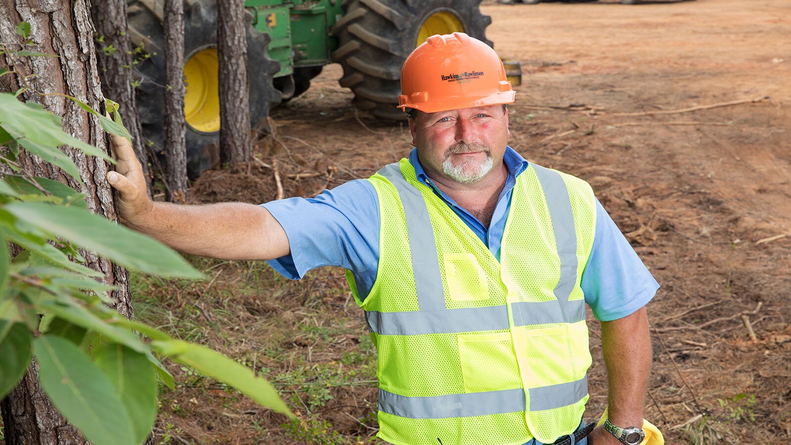 Ray Cobb, in a hard hat and safety vest leans against a tree with his John Deere machines behind him on a logging site in Alabama