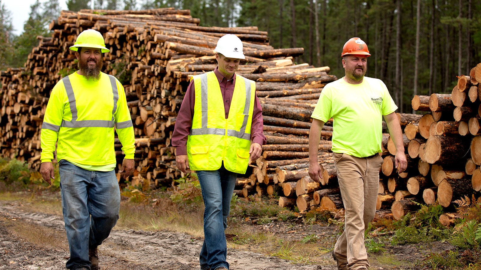 Three men walk in front of a large stack of cut logs.