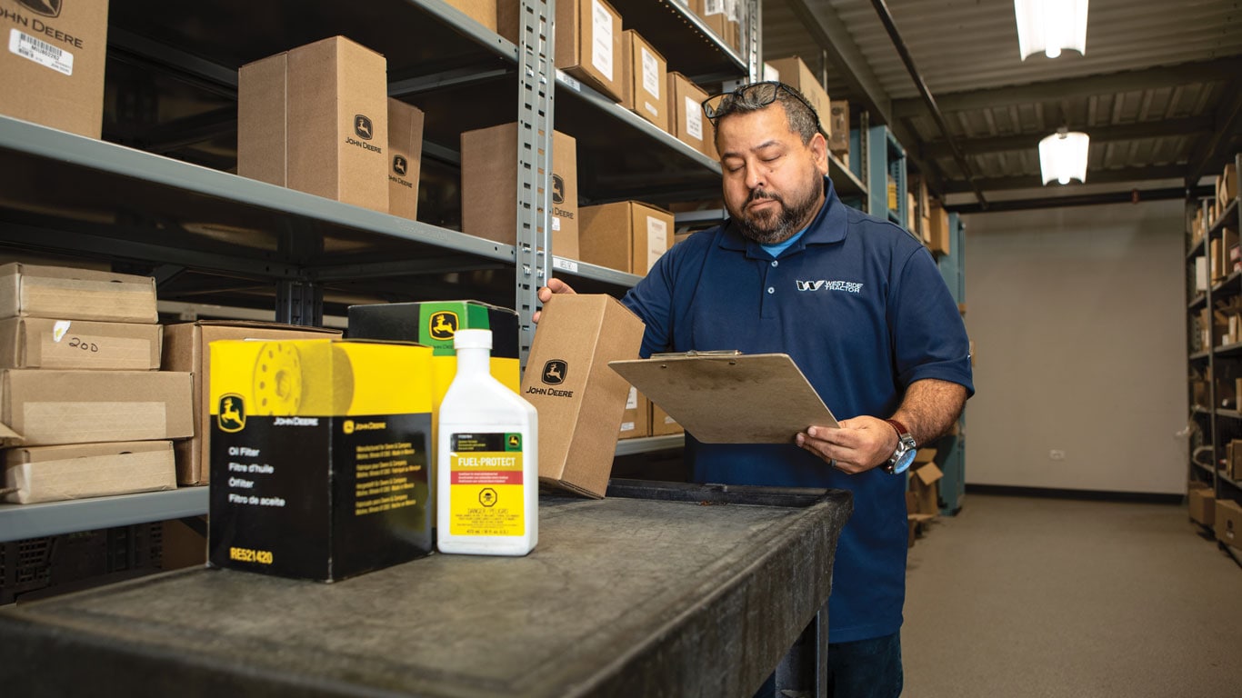 A person in a blue shirt examines a box labeled "John Deere" while holding a clipboard in a storage area filled with equipment.