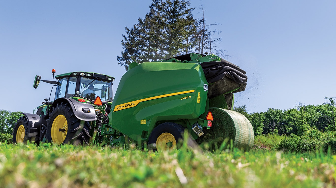 Low-level picture take from the ground with hay blurred in the foreground and the focus of the image on the new John Deere V462M Round Baler being pulled by a John Deere Tractor with a round bale being ejected
