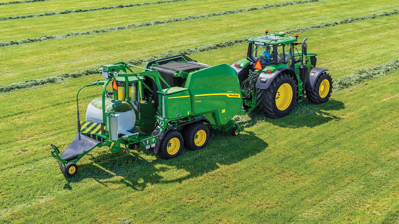 New John Deere V462M Round Baler being pulled by a John Deere 6M Tractor in a green field with multiple windrows in the background and the round baler is wrapping a bale in the back of the baler