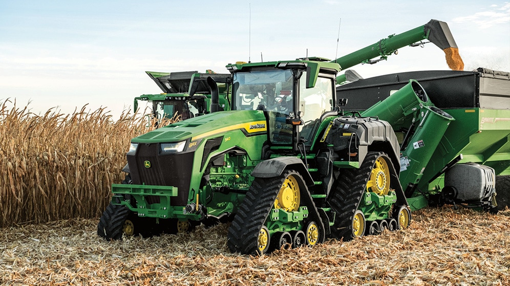 modern John Deere 8rx tractor pulls a storage cart while a harvester transfers harvested wheat