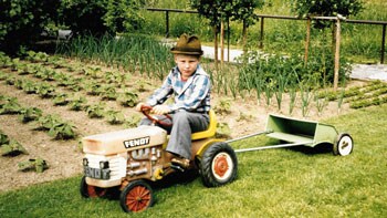 Child driving a toy lawn mower in the grass with garden in background.