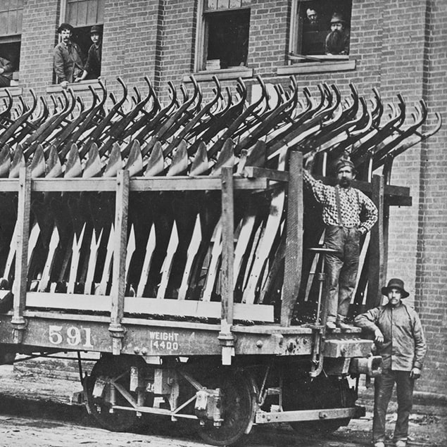Une photographie de 1882 de trois hommes se tenant à côté d'un wagon de Deere & Co chargée de charrues en acier prêtes à être expédiées, avec les employés de l’usine qui regardent par la fenêtre dans l’immeuble derrière